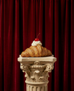 A croissant with cream and a cherry on top stands on an ornate stone pillar in front of a red velvet curtain.