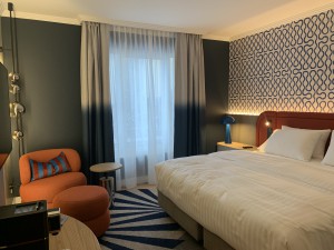 A modern hotel room with a large bed, an orange armchair with an ottoman, a patterned blue carpet, and a window with semi-sheer curtains.