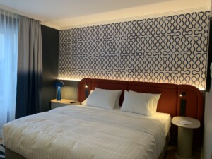Modern hotel bed with a terracotta-colored headboard in front of a wall with a blue and white graphic pattern and integrated lighting.
