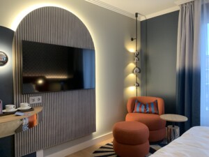A modern hotel room area with a wall-mounted TV, an orange armchair with a matching ottoman, a floor lamp with multiple light bulbs, and a small round side table next to a bed.