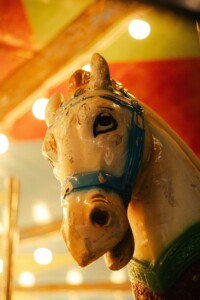 A detailed, painted wooden figure of a horse head with blue bridle against a blurred, warmly lit background.