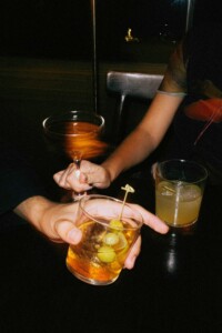 Two people sitting at a dark table, each holding a glass with an alcoholic drink, one of which is garnished with green olives.