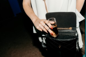 Close-up of a person placing a champagne glass into an open, black quilted bag with a gold chain, their hand wearing several gold rings and red nail polish.