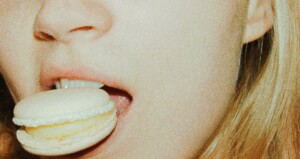 A person with long blonde hair biting into a light-colored macaron.