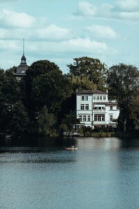 A villa on the shore of a calm Potsdam lake, surrounded by tall trees, while a person paddles on the water in a small boat.