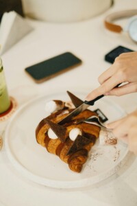 A close-up top-down view of a white plate, where a two-toned chocolate croissant is being cut with a knife and fork. The croissant is decorated with marshmallows, triangular chocolate pieces, and cocoa powder. In the blurred background, a smartphone and headphones rest on a light-colored table.