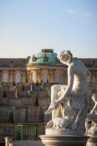 A white marble sculpture of a seated person holding their foot stands in front of the vineyard terraces and Sanssouci Palace.