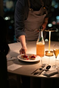 A person in an apron serving two small desserts on a white plate in a dimly lit restaurant, while a bottle and two glasses with drinks stand on the table.