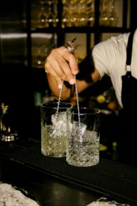 A bartender holding two long stirring rods inside two ice-filled decorative glasses on the counter.