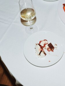 A top-down view of a white plate on a white tablecloth. On the plate are the remains of a dessert with cream, berries, and dark cake. Next to it stands an elegant wine glass, partially filled with white wine.