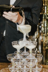 A person in a dark suit pouring champagne into a pyramid structure of multiple champagne glasses, with the foam overflowing.