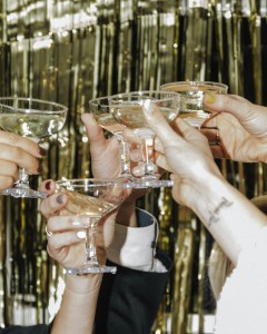 Hands clinking champagne glasses in front of a golden glitter curtain.
