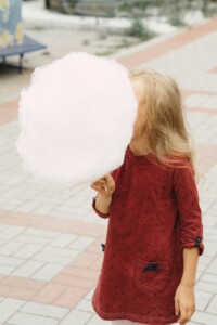 A child with long blonde hair wearing a red polka dot dress and holding a large cloud of cotton candy in front of their face.